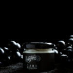 Unmistakable - Vegane Premium Tattoopflege - Care Moisture Balm Glas auf schwarzem Sand zwischen schwarzen Weintrauben
