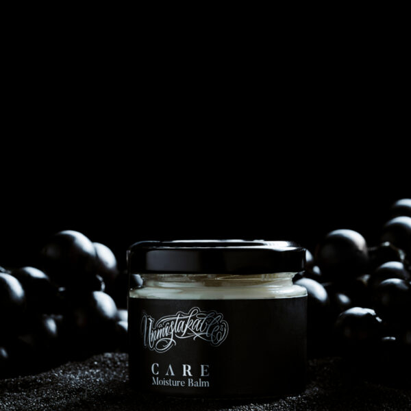 Unmistakable - Vegane Premium Tattoopflege - Care Moisture Balm Glas auf schwarzem Sand zwischen schwarzen Weintrauben