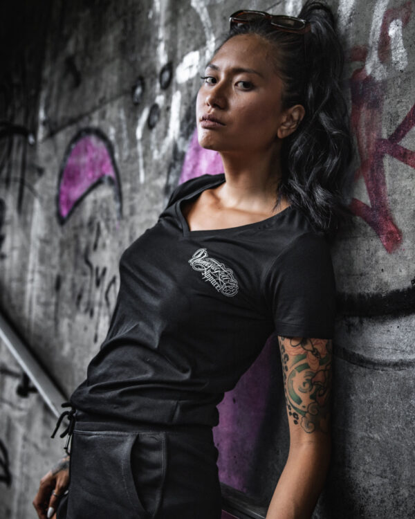 Unmistakable - V-Neck T-Shirt - Junge Frau mit dunklen Haaren lehnt an Graffiti-Wand